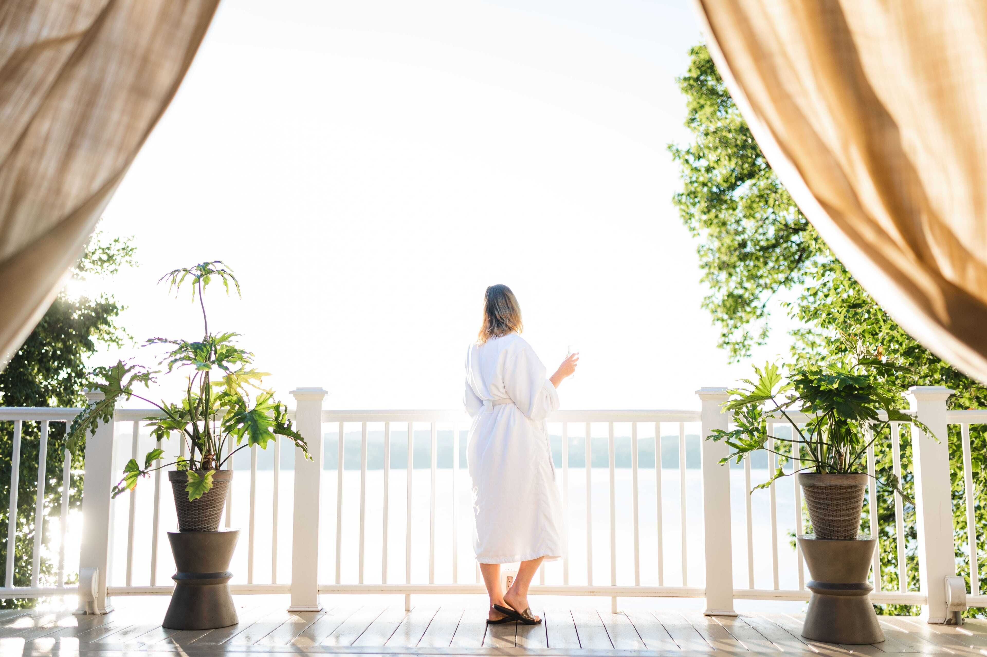 A person in a white robe standing on a wooden deck with potted plants, overlooking a white railing and a body of water.