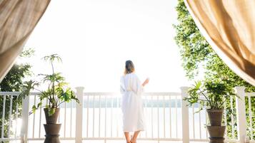 A person in a white robe standing on a wooden deck with potted plants, overlooking a white railing and a body of water.