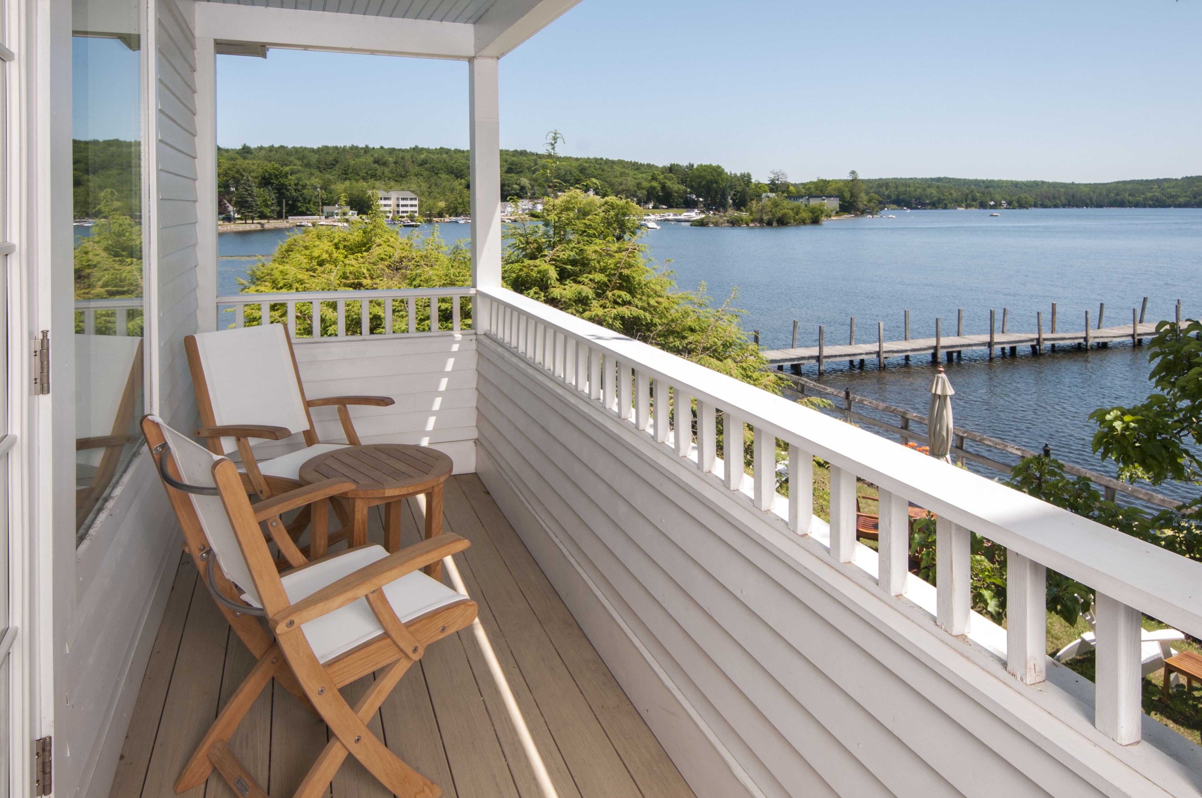 A wooden deck with a white railing overlooking a lake, a wooden table with chairs, and a clear view of the water and distant shoreline.