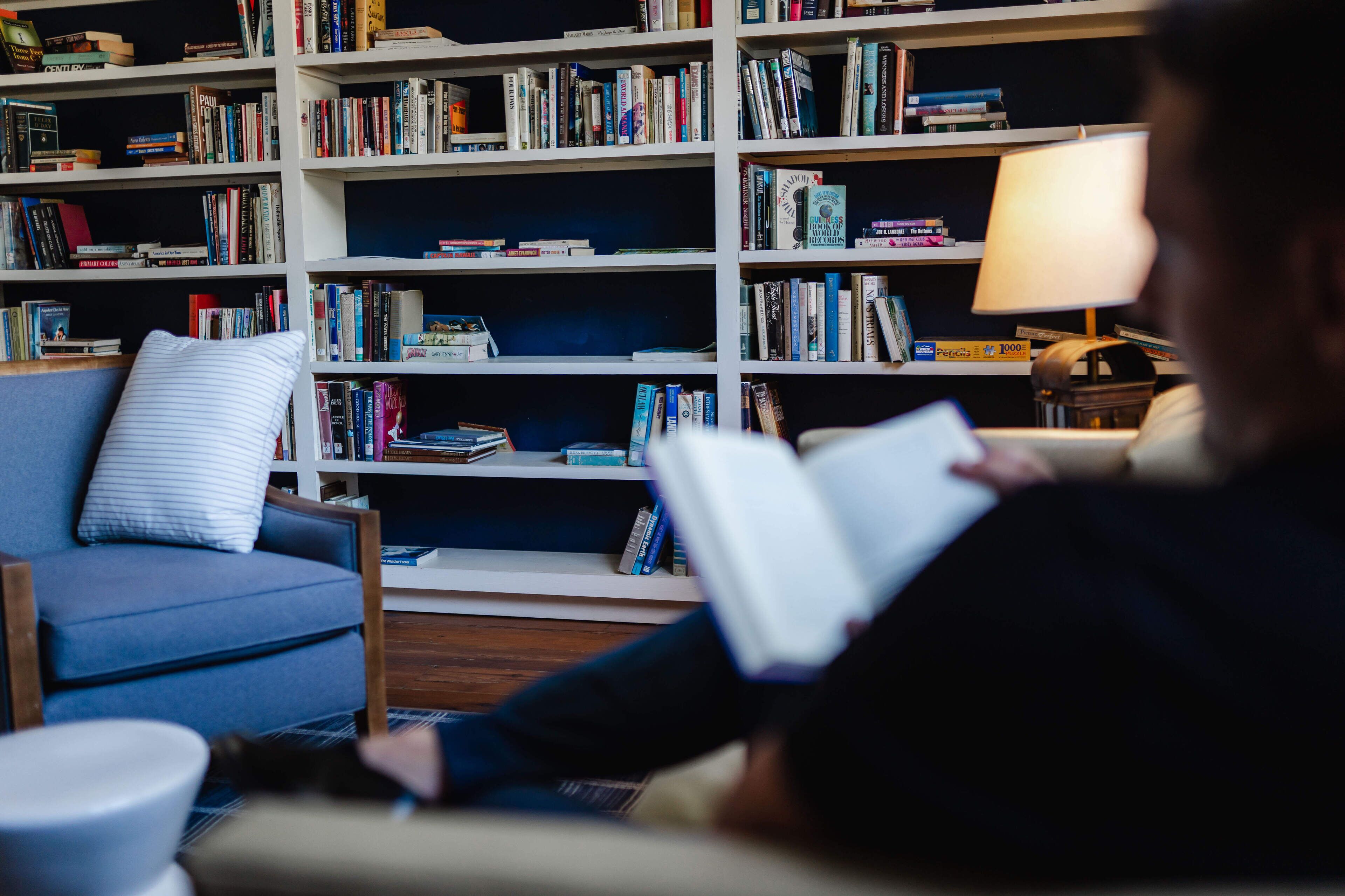 A cozy reading nook with a blue armchair, a white pillow, and a bookshelf filled with books.