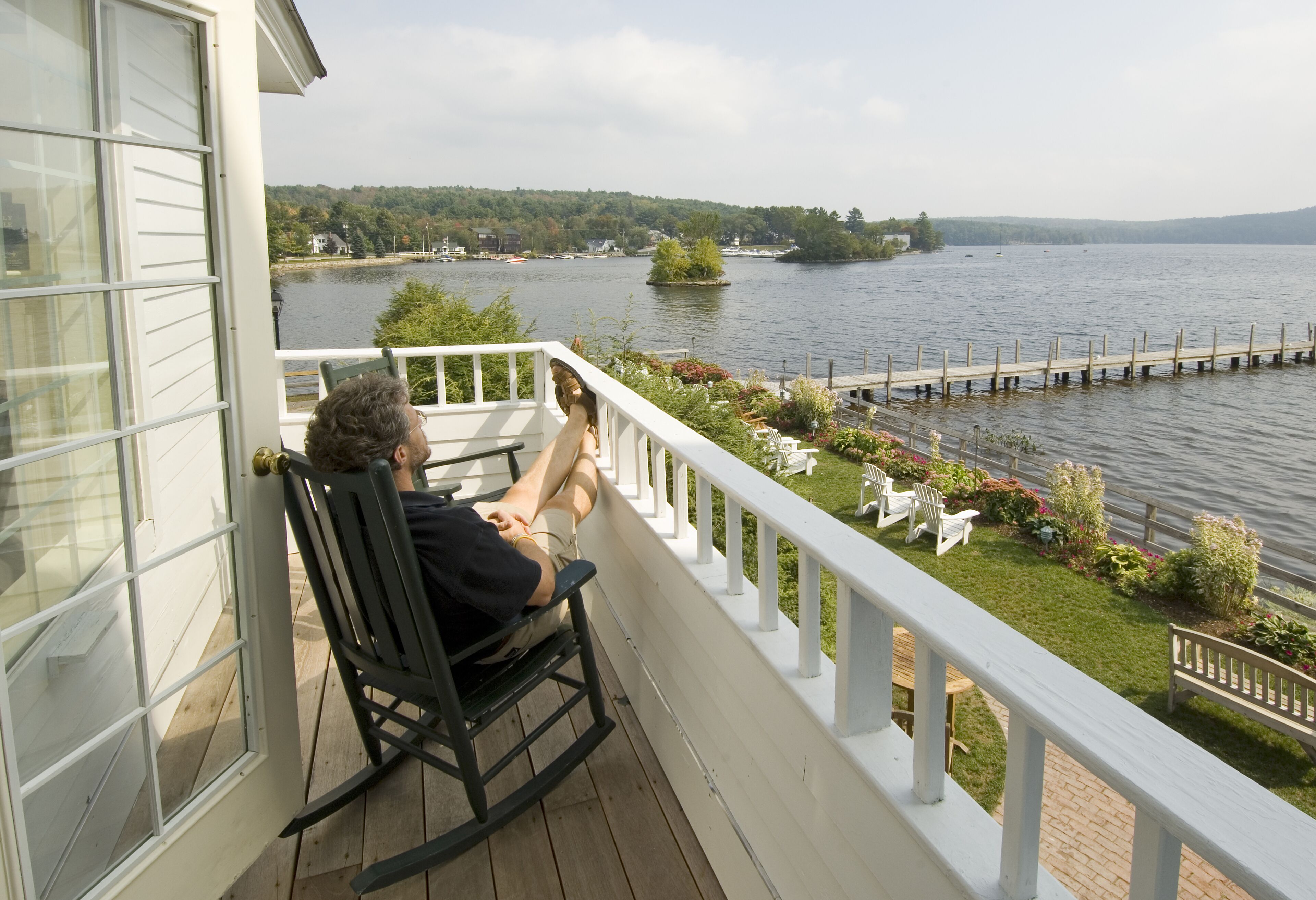 A person sitting on a rocking chair on a wooden deck with a white railing, overlooking a lake with a dock and greenery.