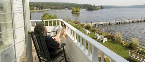 A person sitting on a rocking chair on a wooden deck with a white railing, overlooking a lake with a dock and greenery.
