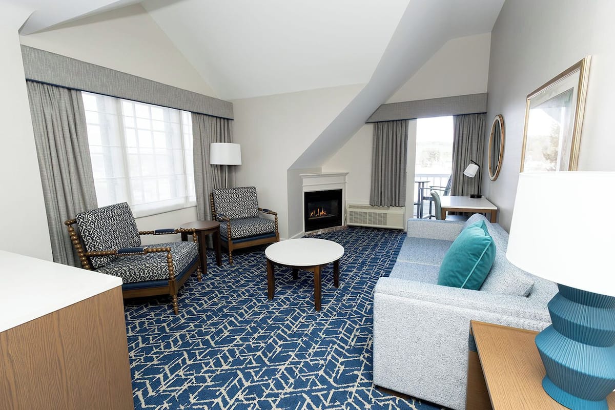 A hotel room with a fireplace, a sofa, two armchairs, a round table, and a patterned carpet.