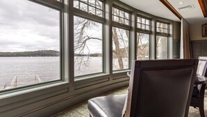 Habitación con amplios ventanales que ofrecen vistas a un lago y colinas. Se observa un sillón de cuero y una mesa con lámpara.