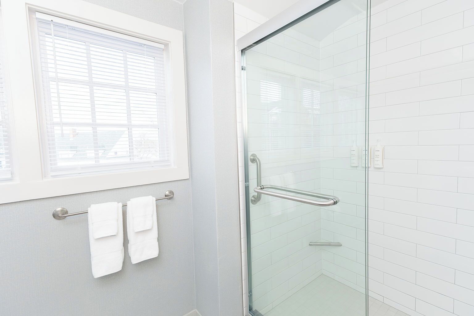 Een moderne badkamer met een glazen douchedeur, witte betegelde wanden en een raam met zonwering.