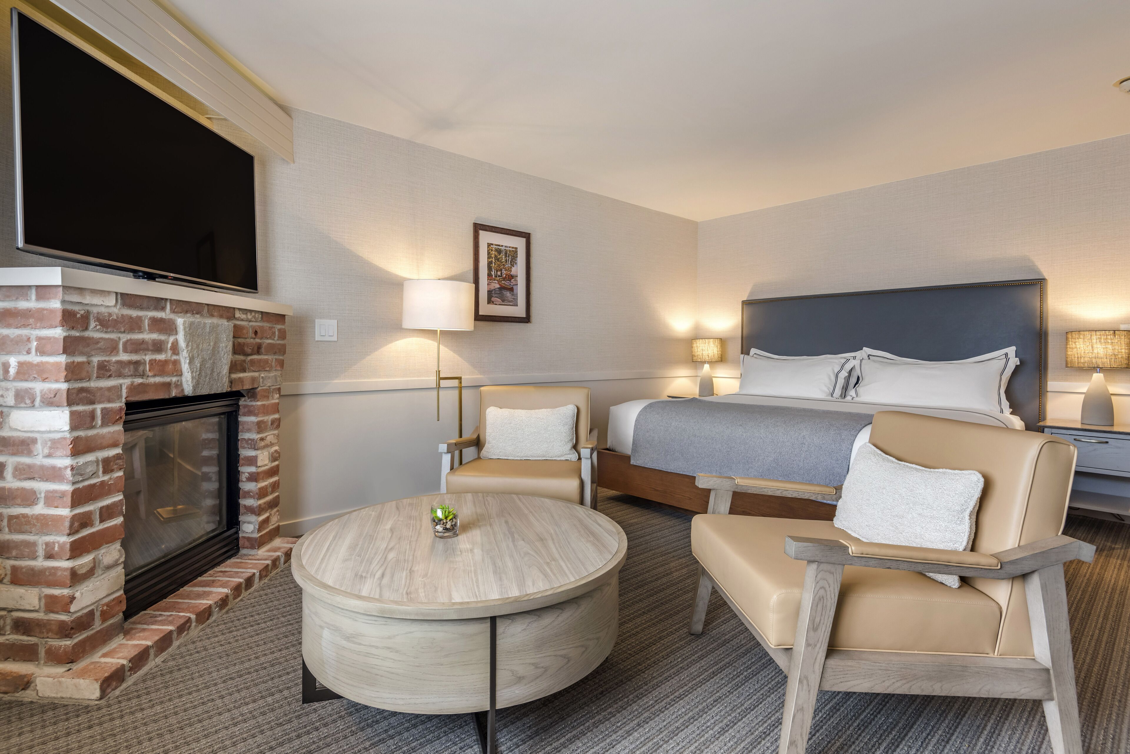 Ein Hotelzimmer mit Kamin aus Backsteinen, einem runden Holztisch, zwei Sesseln, einem Bett mit Kopfteil und einem flachbildfernseher an der Wand.