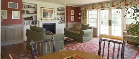 A cozy living room with two green sofas, a wooden coffee table with a chessboard, a fireplace, and a large window with an exit sign.