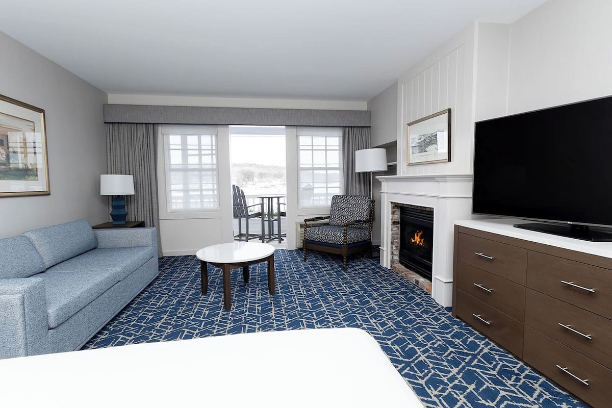 A hotel room with a blue patterned carpet, a sofa, a small table, a chair, a fireplace, a TV, and a window with curtains.
