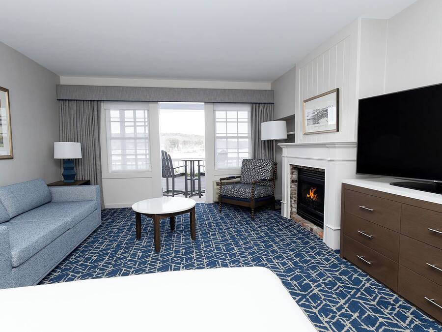 Habitación de hotel con alfombra azul estampada, sofá, mesita, silla, chimenea, televisor y ventana con cortinas.