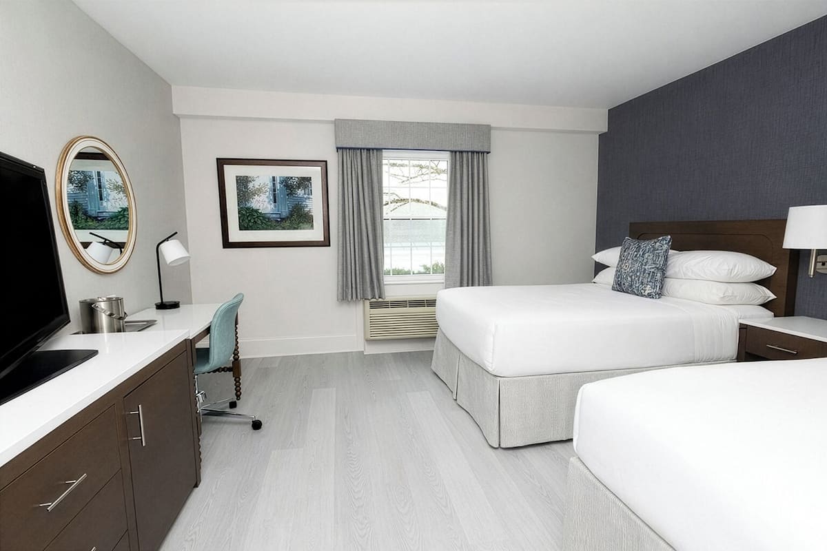 A hotel room with a large bed, a smaller bed, a desk with a chair, a TV, and a window with curtains.