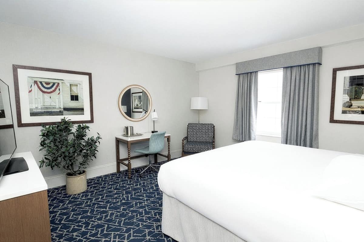 A hotel room with a large bed, a desk with a chair, a small table with a lamp, a potted plant, and a window with curtains.
