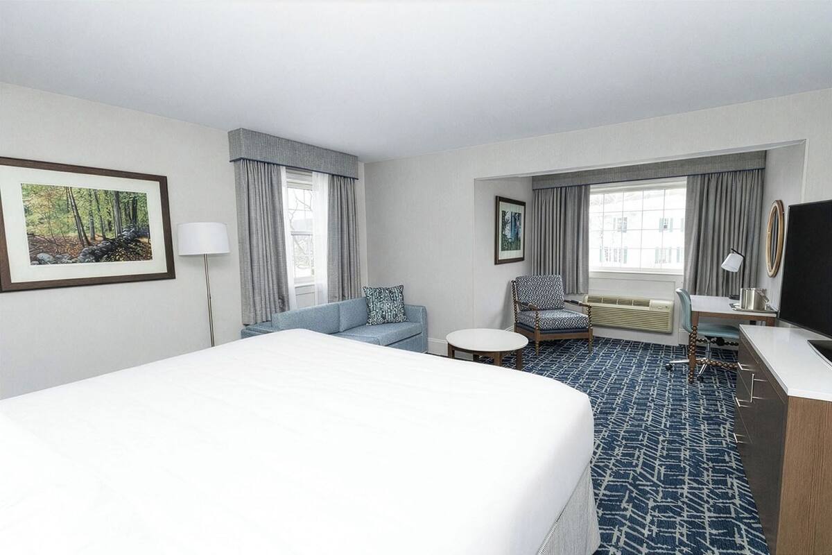 A hotel room with a large bed, a sofa, a desk, and a chair. There are two windows with curtains, a painting on the wall, and a lamp.