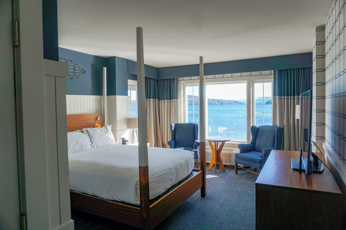 A hotel room with a four-poster bed, a desk, two chairs, a small table, and a view of the water.
