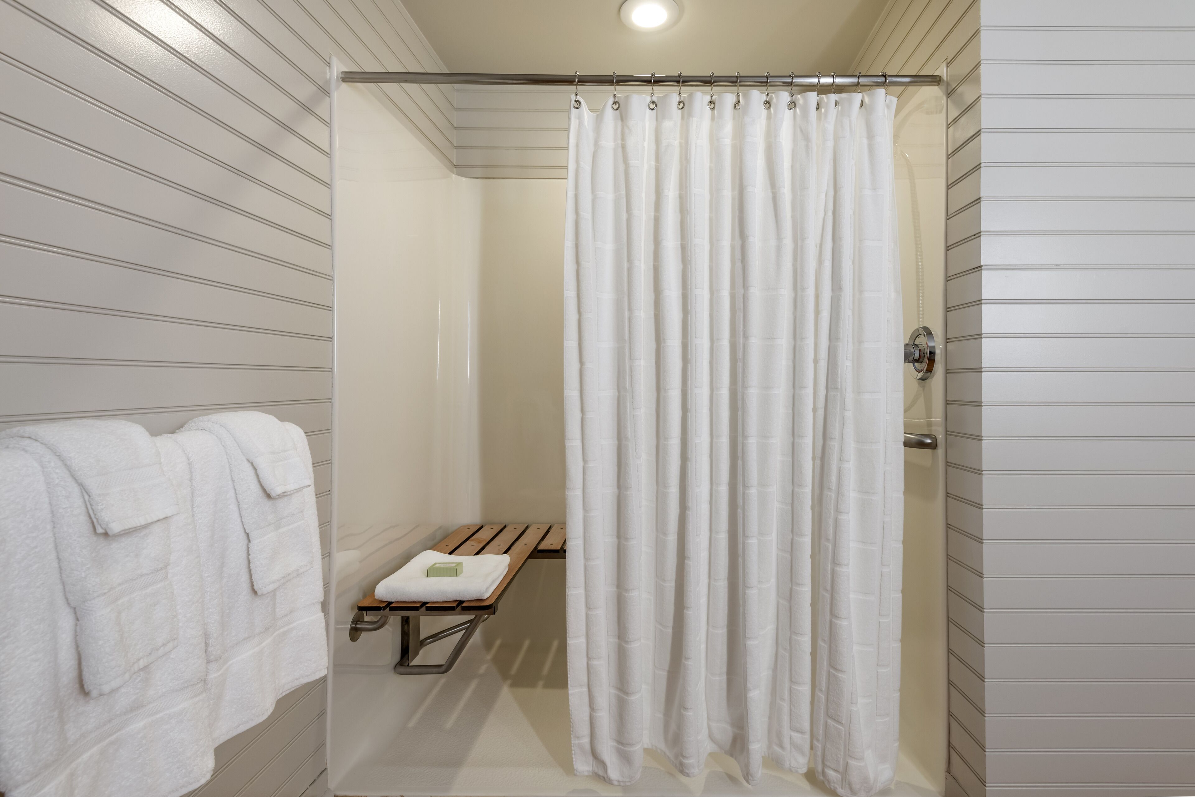 A shower with a white curtain, a bench, and a towel rack with towels.