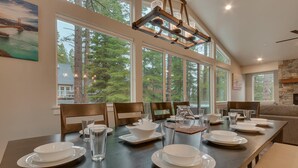 A dining area with a long table set for six, large windows, and a stone fireplace. - Golden Tee, Classic Arcade, Slot Machine, Spa, 7mins to Beach/ Lake, Newly Built – Sierra Escape (South Lake Tahoe)