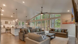 A spacious living area with a large window, a ceiling fan, and a kitchen in the background. - Golden Tee, Classic Arcade, Slot Machine, Spa, 7mins to Beach/ Lake, Newly Built – Sierra Escape (South Lake Tahoe)