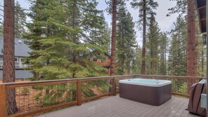 A wooden deck with a hot tub, surrounded by tall pine trees and a house in the background. - Golden Tee, Classic Arcade, Slot Machine, Spa, 7mins to Beach/ Lake, Newly Built – Sierra Escape (South Lake Tahoe)