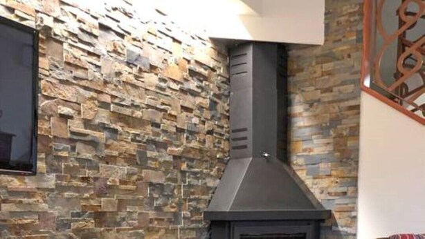 Ein gemütliches Zimmer mit einer Steinwand, einem schwarzen Kamin, einem Korbstuhl mit Kerzen und einem Korbtisch.