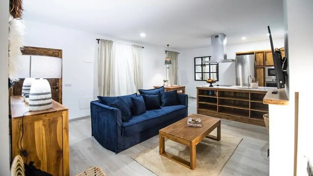 Ein modernes Wohnzimmer mit einem blauen Sofa, einem Holztisch und einer Küchenzeile mit Holzschränken und einem Einbauofen.