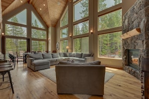 A spacious living room with a large sectional sofa, a fireplace, and large windows offering a view of the forest.