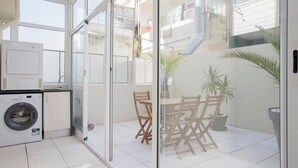 Ett tvättrum med tvättmaskin och torktumlare, en glasdörr som leder ut till en balkong med ett bord och stolar, och utsikt över den yttre delen av byggnaden.