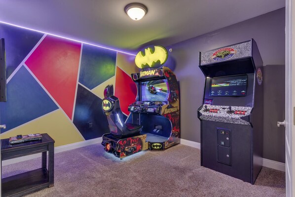 Ein Spielbereich mit zwei Batman-Automaten, einem kleinen Tisch mit Büchern und einem farbenfrohen, geometrischen Wanddesign.