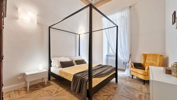 Un dormitorio con una cama con dosel, una mesita de noche, una silla y una ventana con cortinas.