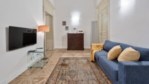 Una sala de estar con una televisión de pantalla plana, una mesa de centro de vidrio, un sofá azul con cojines, una alfombra estampada y un mueble de madera.