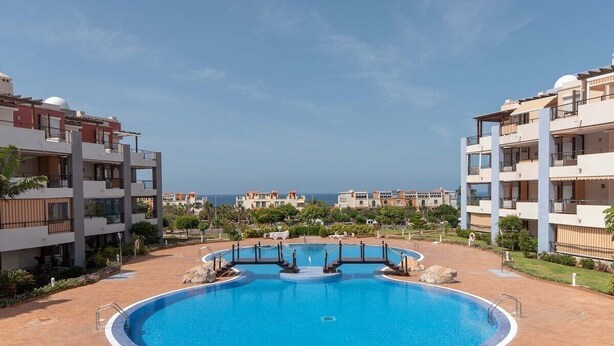 A swimming pool surrounded by modern buildings with balconies, a clear sky, and a distant view of the sea. - El Rincon 3 (Los Cristianos)