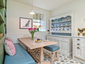 A kitchen with a wooden table, a bench, a vase with flowers, and a cabinet with plates. - Home Farm Cottage - UK10382 (Glenkindie, near Alford)