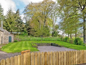 A stone building with a green door, a wooden fence, and a sign that reads 'HOME FARM' in a lush green landscape with trees and a gravel path. - Home Farm Cottage - UK10382 (Glenkindie, near Alford)