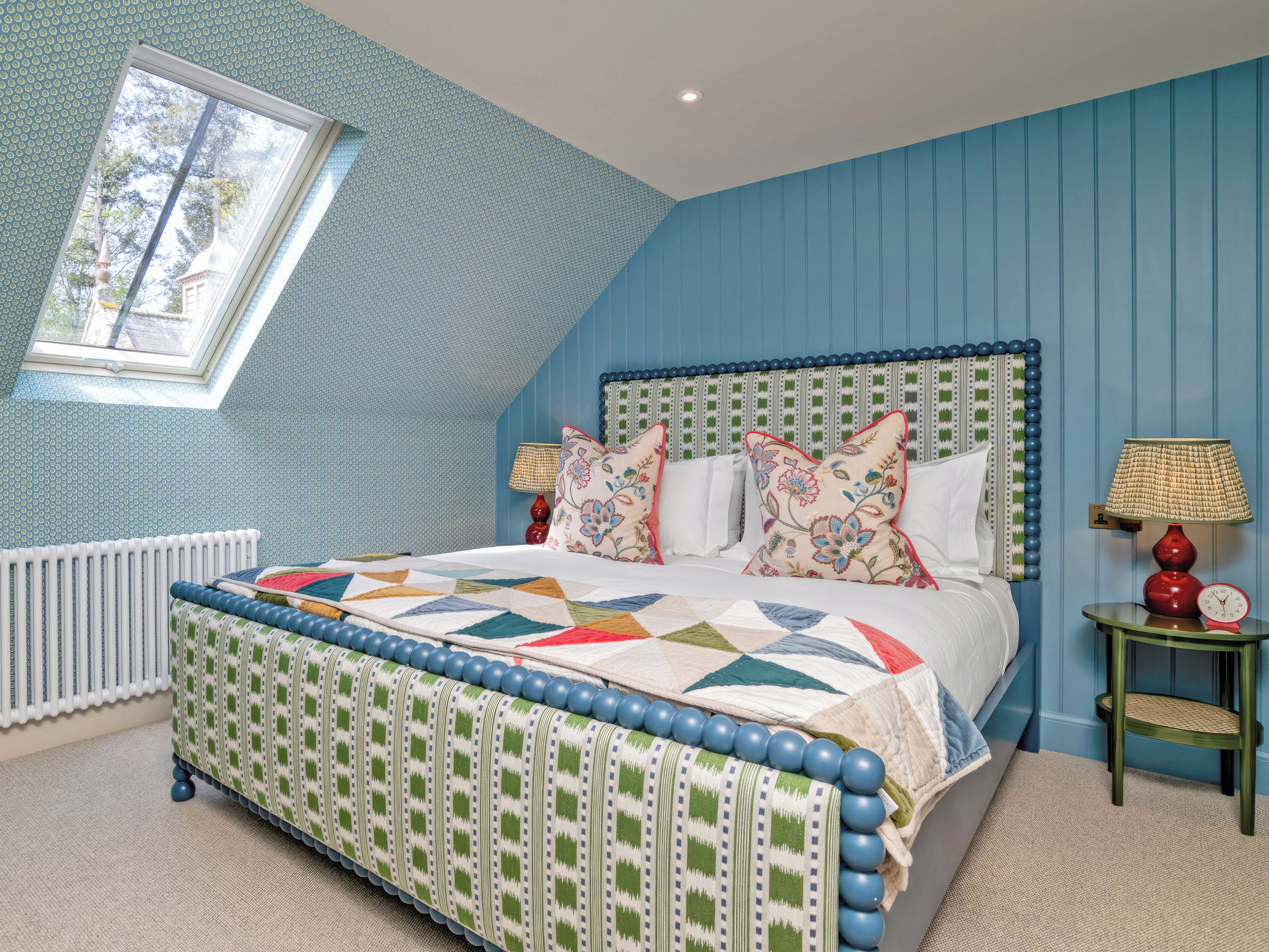 A bedroom with a sloped ceiling, a bed with a patterned bedspread, decorative pillows, a bedside lamp, and a clock on a nightstand.