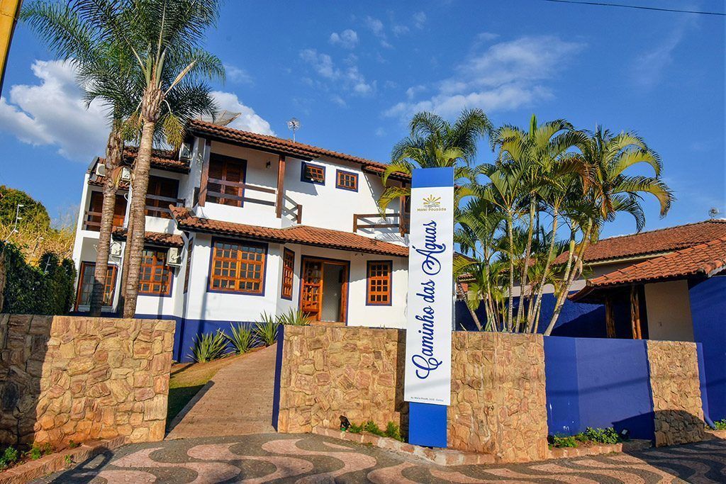 A two-story building with a tiled roof, a stone wall entrance, and a sign reading "Camino das Águas."