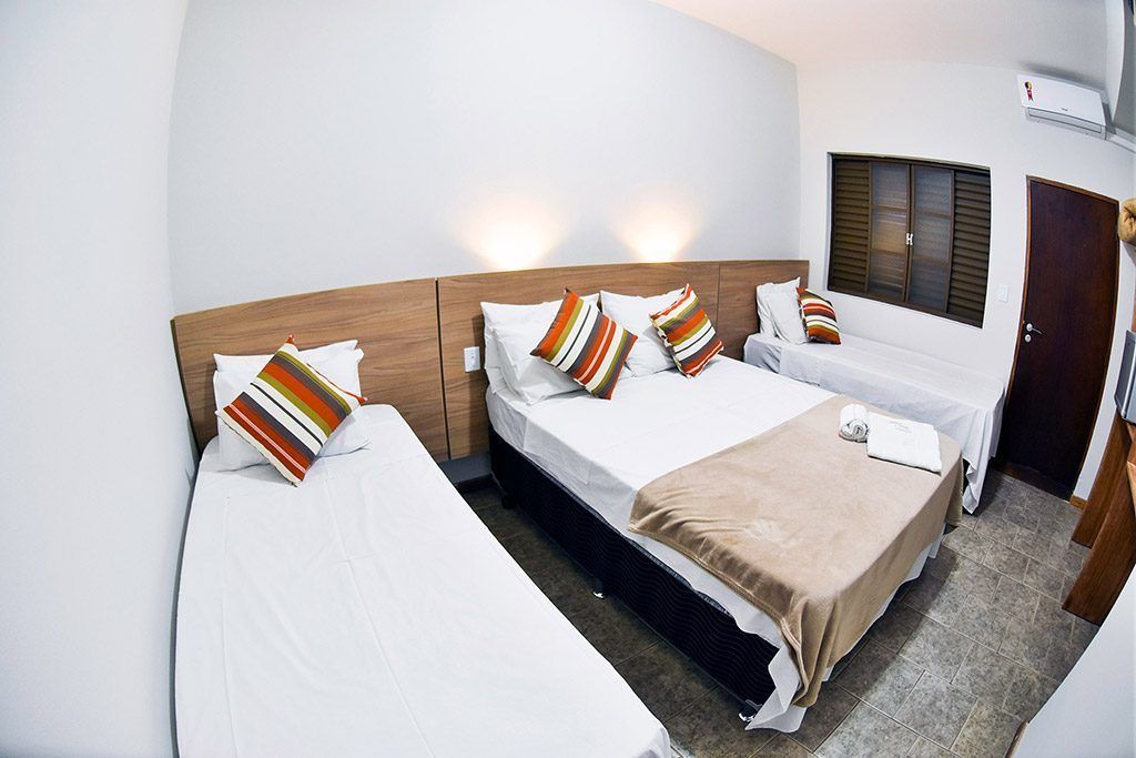 Une chambre d’hôtel avec trois lits, chacun doté de draps blancs et d’oreillers à rayures. On y trouve une tête de lit en bois, une fenêtre avec des stores et une porte.