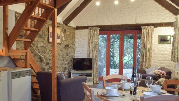 A cozy room with a stone wall, wooden staircase, a television, a dining table set for four, and a sofa. - 2 bedroom accommodation in Houghton, near Haverfordwest (Houghton, near Haverfordwest)