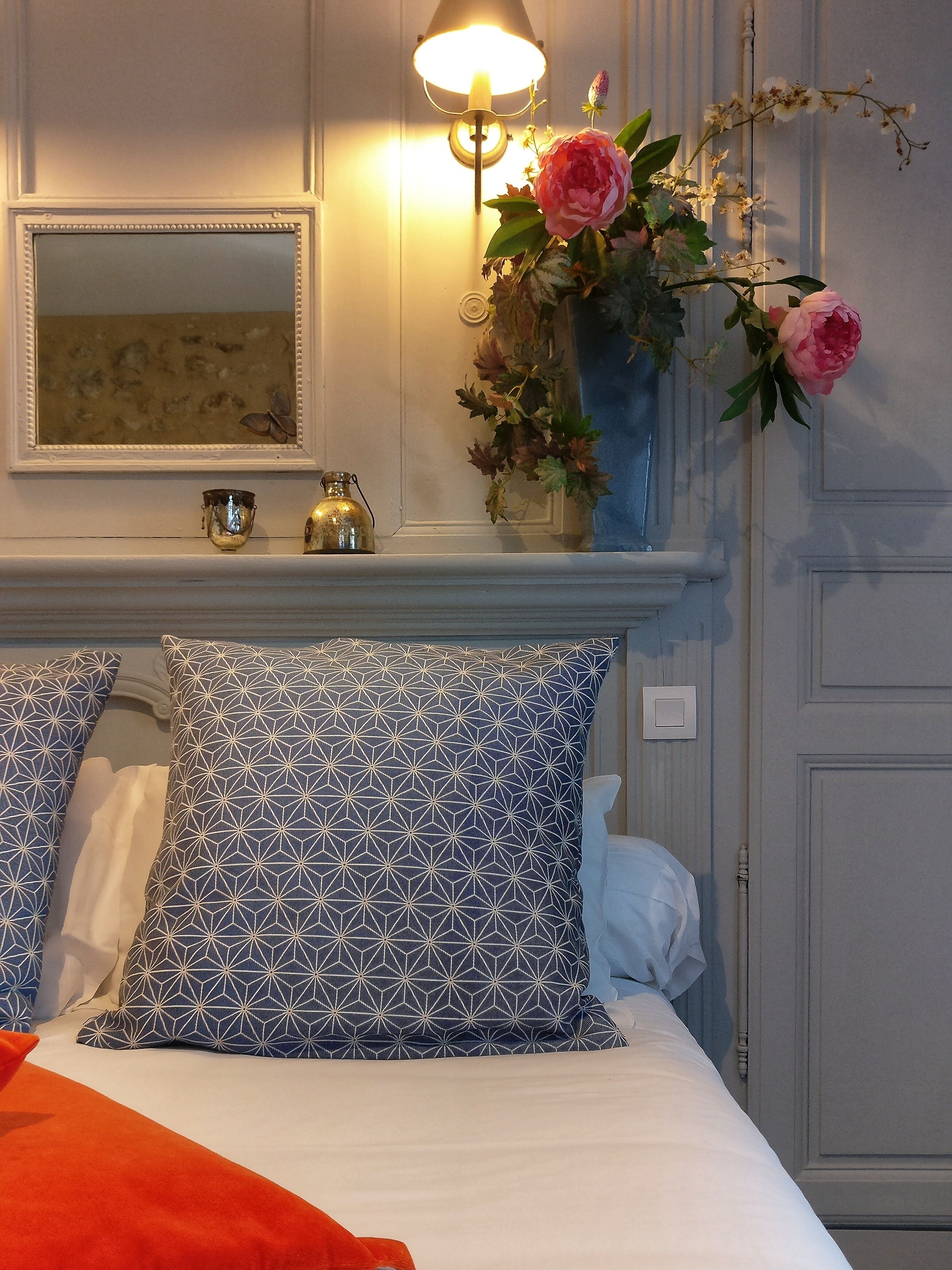 A bedroom with a bed, pillows, a vase with flowers, a wall-mounted lamp, and a mirror.
