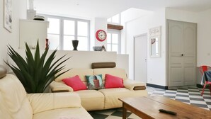 A living room with a beige sofa, a large plant, and a wooden coffee table. There are two clocks on the wall, a door, and a checkered floor. - Notre Dame Chauvigny - Chambres d'hôtes (Chauvigny)
