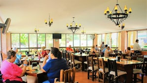 Un restaurante espacioso con numerosas mesas y sillas, ventanas amplias y candelabros colgantes.