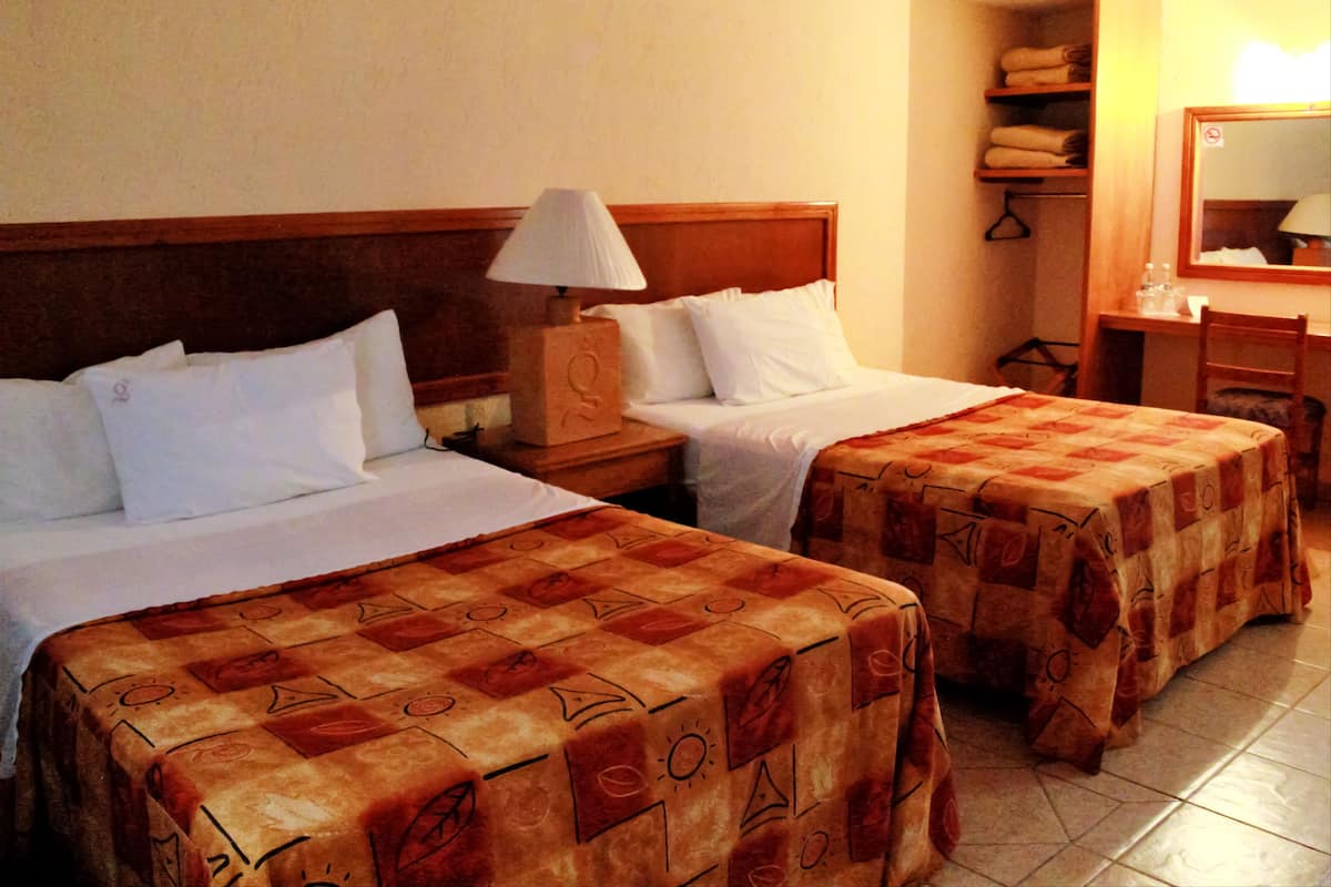 A hotel room with two beds, a nightstand with a lamp, a wooden headboard, a mirror, a chair, and a shelf with towels.