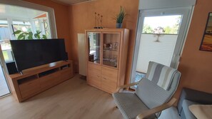 Una sala de estar con un televisor sobre un mueble de madera, un aparador con puertas de cristal, un sillón a rayas y una ventana con persianas.