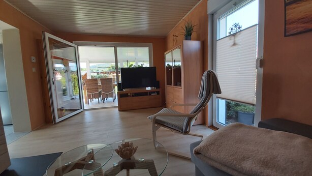 A living room with a TV, a glass coffee table, a cushioned bench, and a sliding glass door leading to an outdoor area with chairs and a table. - Holiday home "Zur Lehns" with garden (Darlingerode)