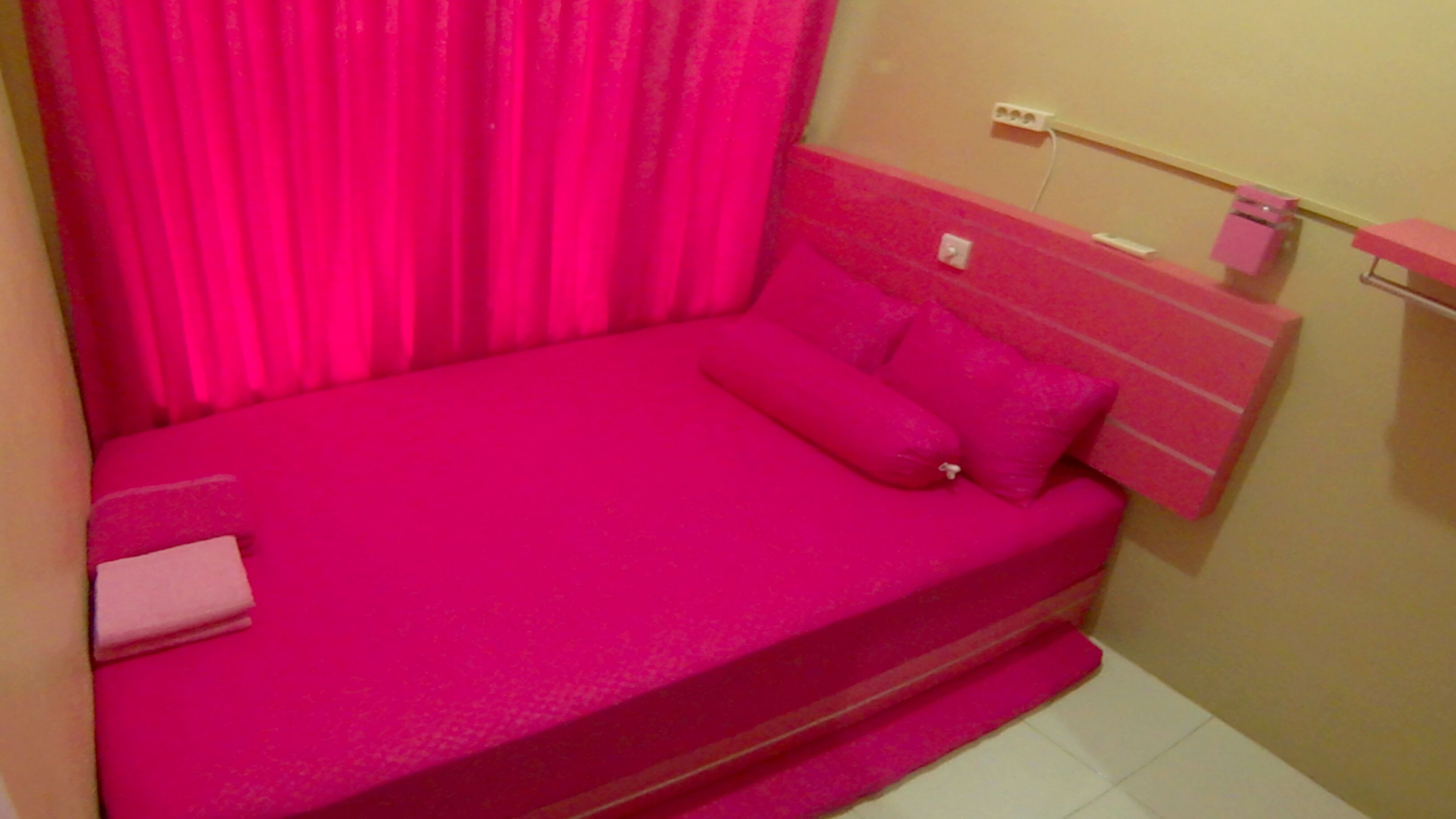 A small, pink-colored room with a bed, a bedside table, and a wall-mounted shelf.