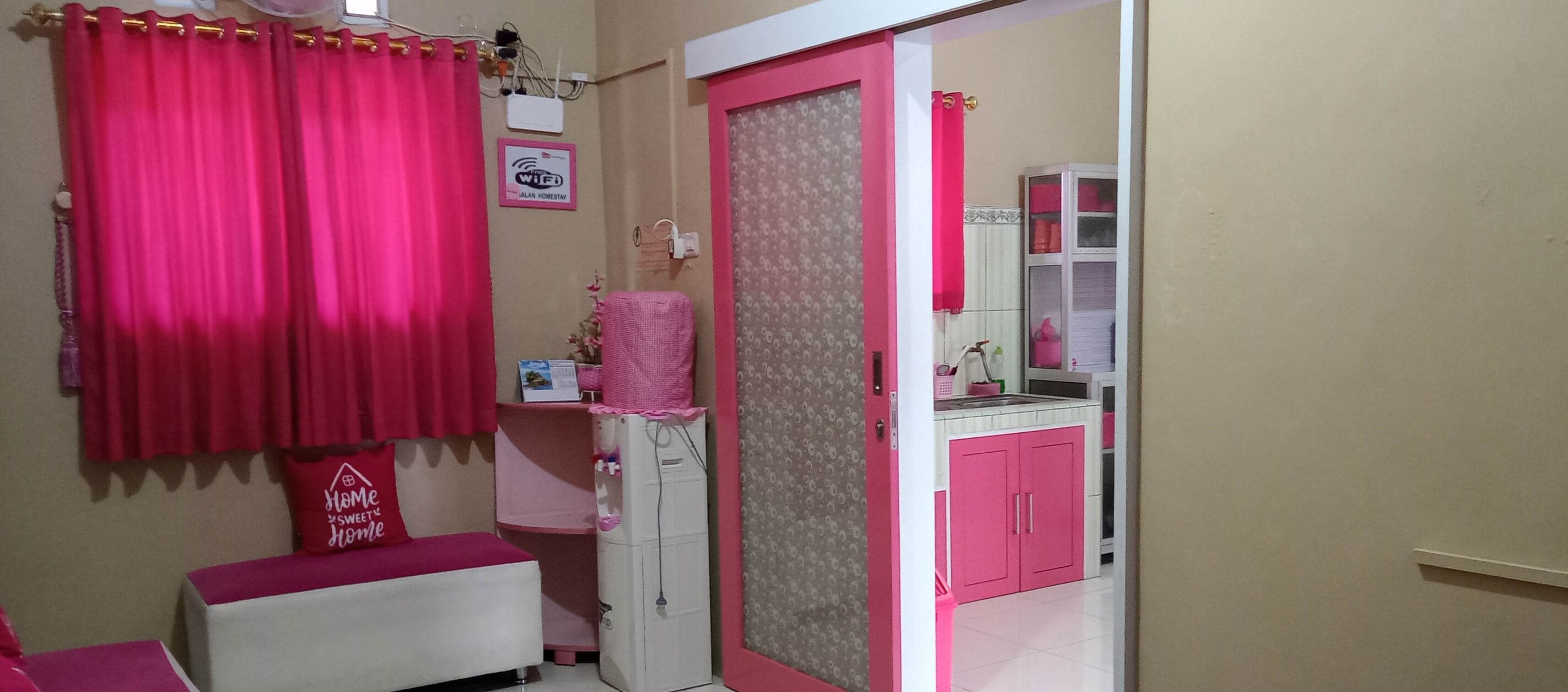 A room with pink curtains, a bed, a small table, a cabinet, and a mirror.