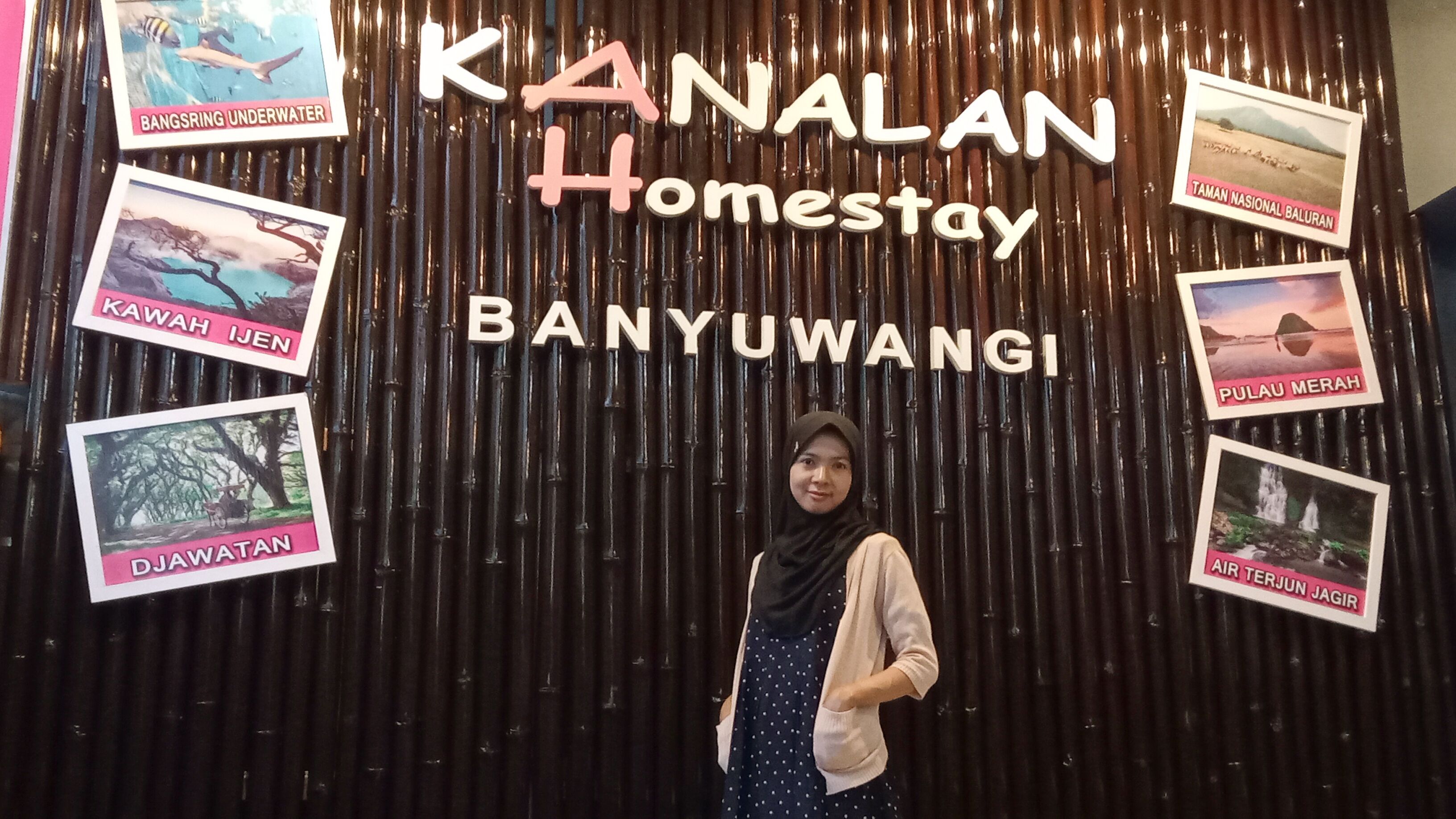 A woman standing in front of a sign for Kanalan Homestay Banyuwangi with photos of local attractions.
