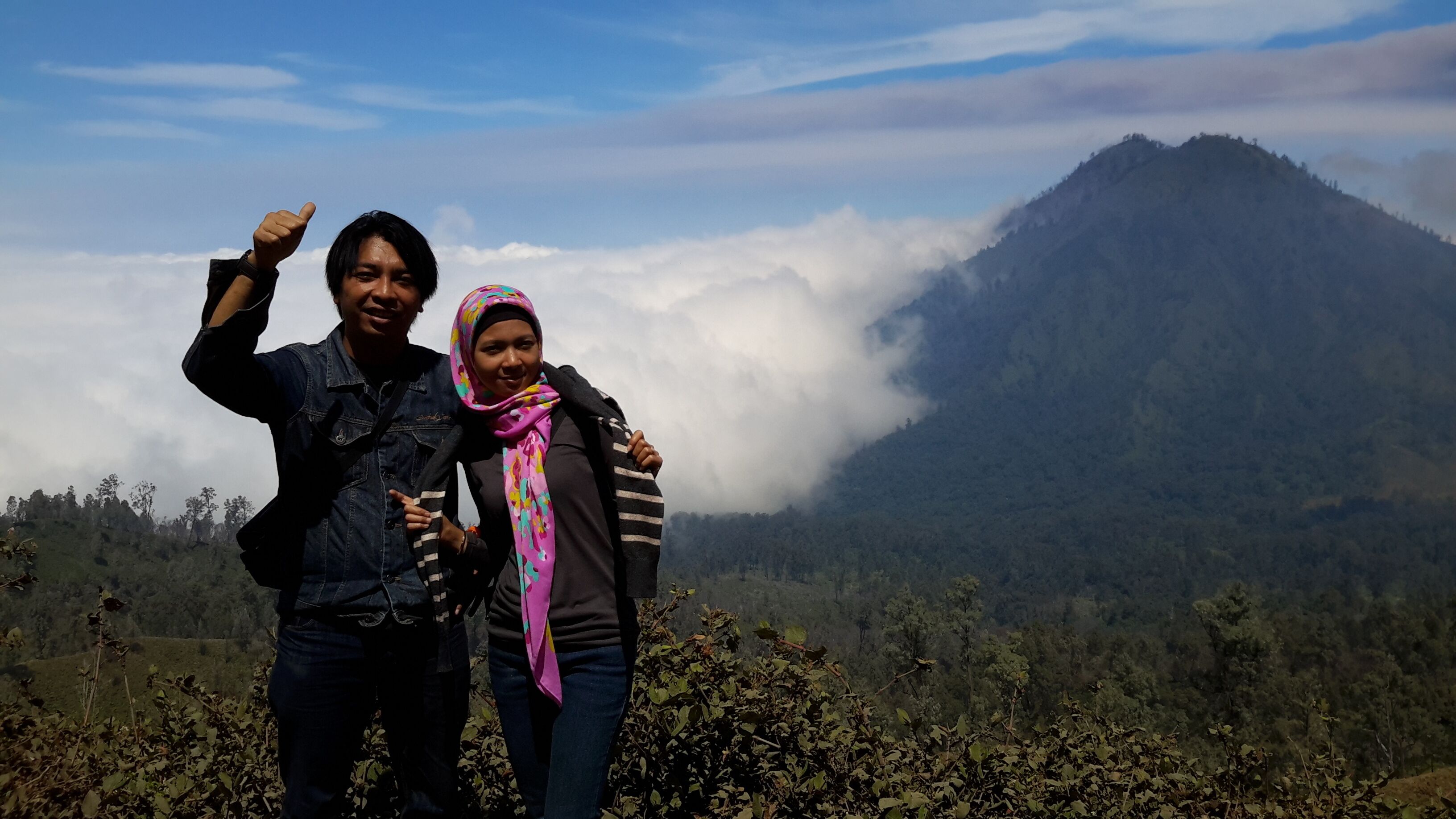 Dos personas de pie en una montaña, con un bosque al fondo y nubes.