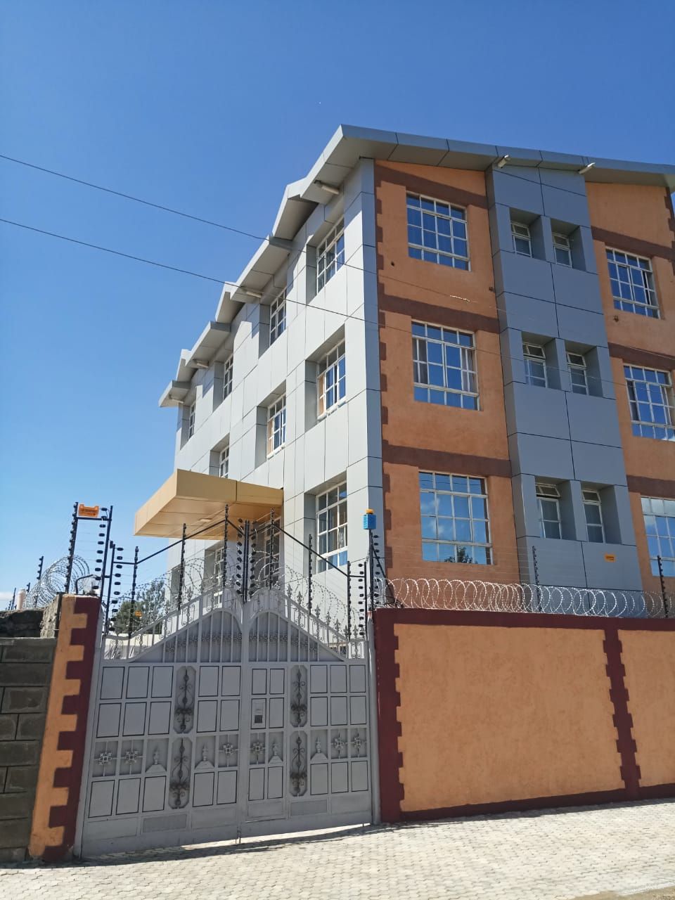 Un edificio de varios pisos con una entrada protegida por un portón, cielo despejado y una línea de electricidad visible.