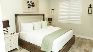 A neatly arranged bedroom with a large bed, a bedside table with a lamp, a world map on the wall, and a window with blinds.