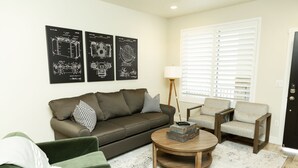 A living room with a green sofa, a round coffee table, and two armchairs. There are three black and white framed posters on the wall.