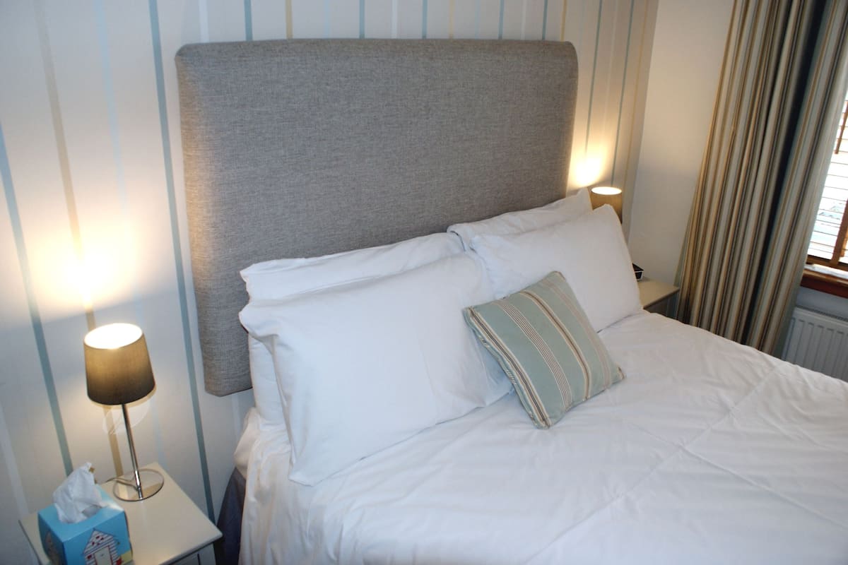 Un lit bien fait, avec du linge de lit blanc et une tête de lit grise, deux lampes de chevet et un coussin à rayures.