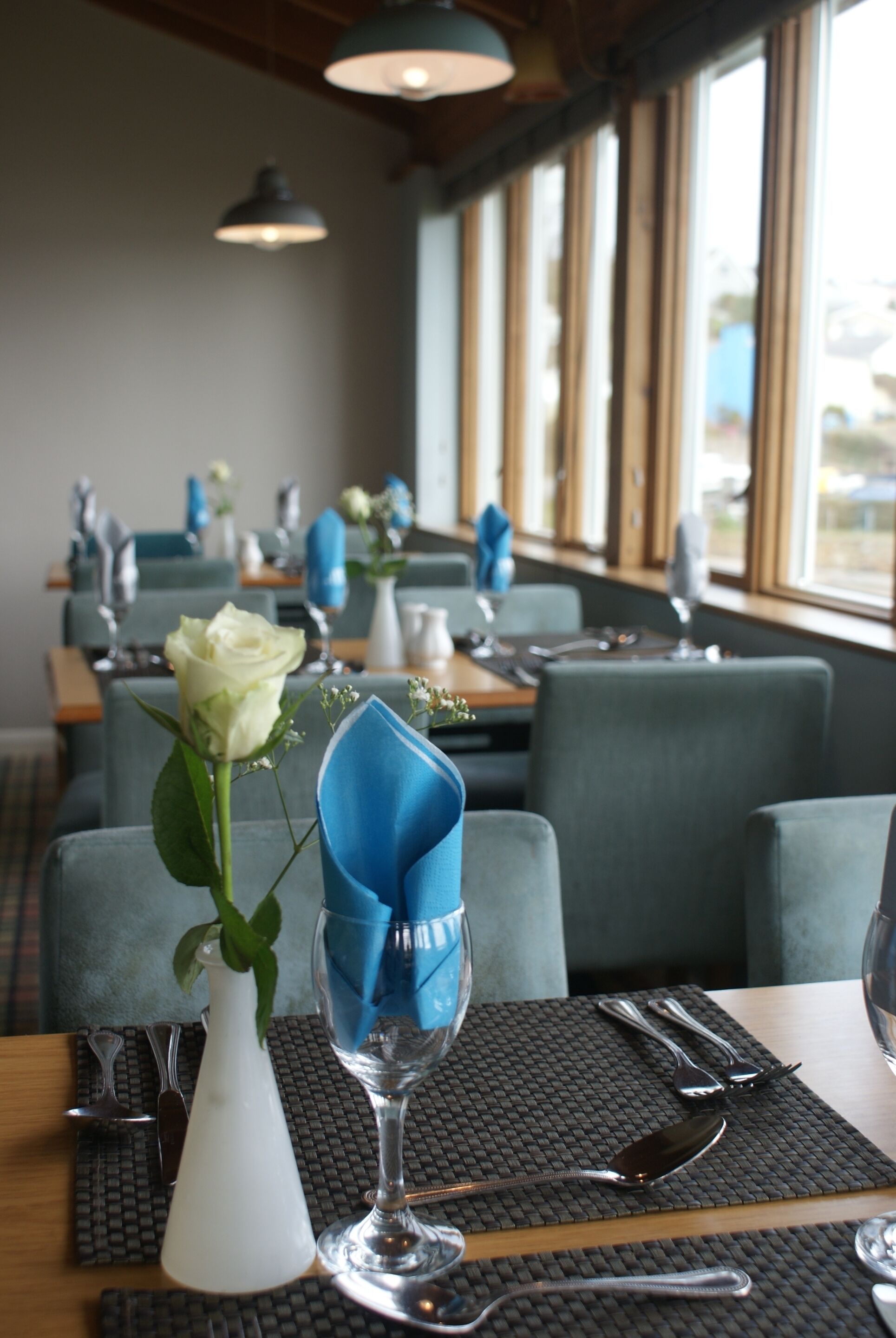 A restaurant interior with tables set for dining, featuring blue napkins, white vases, and a single white rose.